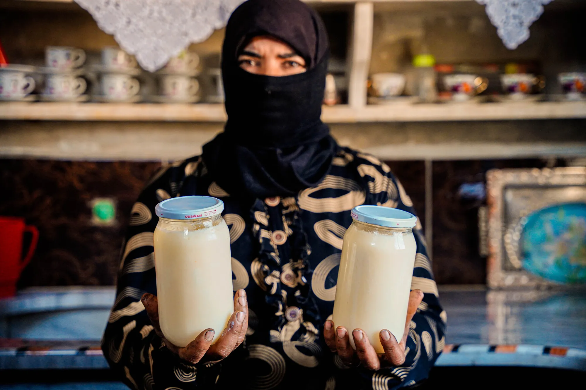 A Syrian woman, her face mostly covered by a black headscarf, holds two jars of a creamy liquid.