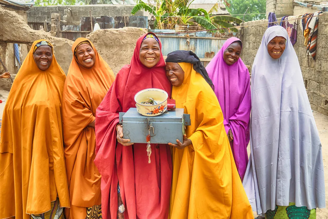 Six joyful VSLA women from Nigeria stand outside laughing in brightly colored robes. Two hold the Village Savings and Loan Association (VSLA) cash box used to secure their shared savings.