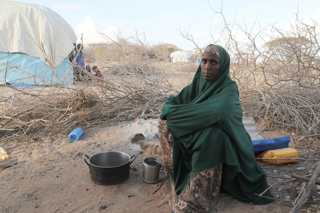 A Somali woman wearing a green shawl and patterned dress sits on the ground in a dry, sparse area surrounded by leafless branches, with an empty cooking pot and metal cup in front of her and dome-shaped tents in the background.
