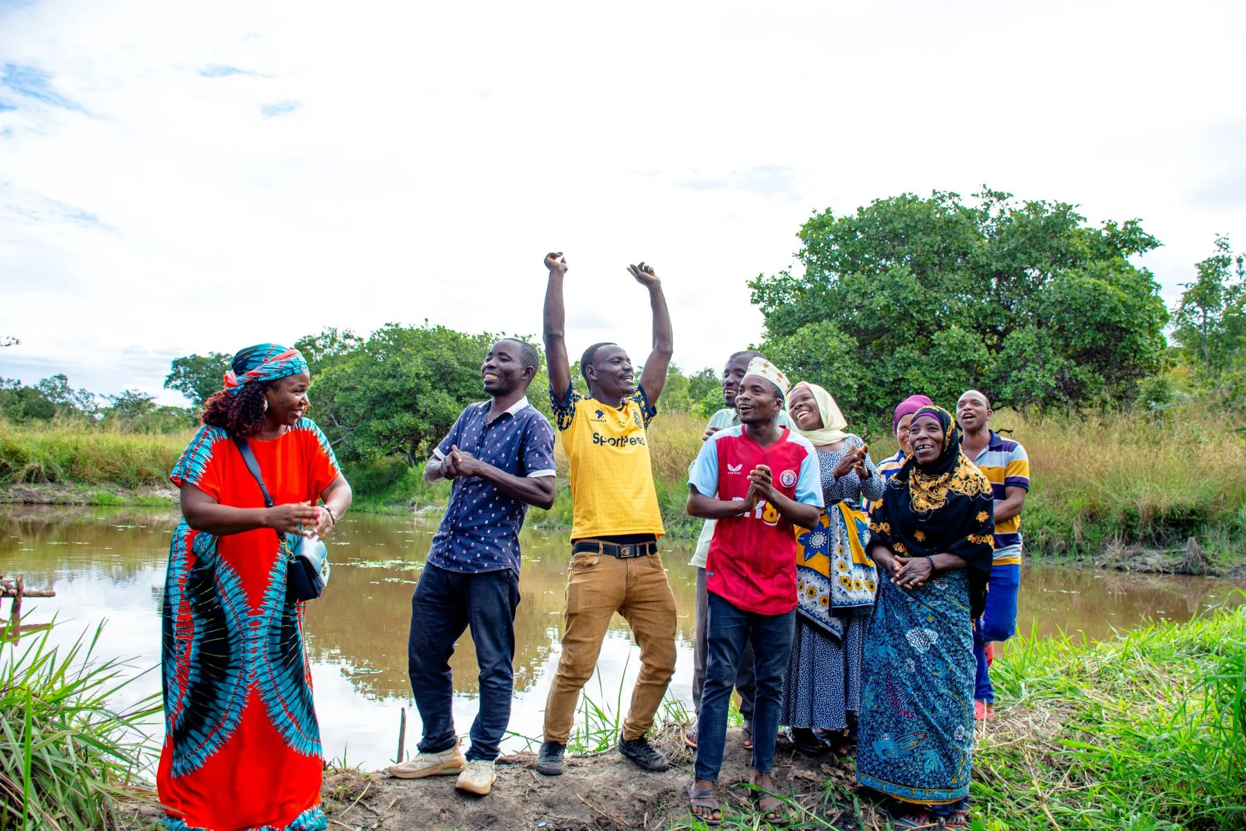 A joyous group of men and women, members of a Village Savings and Loan Association (VSLA) in Tanzania, stand cheering by a body of water with lush greenery behind them.