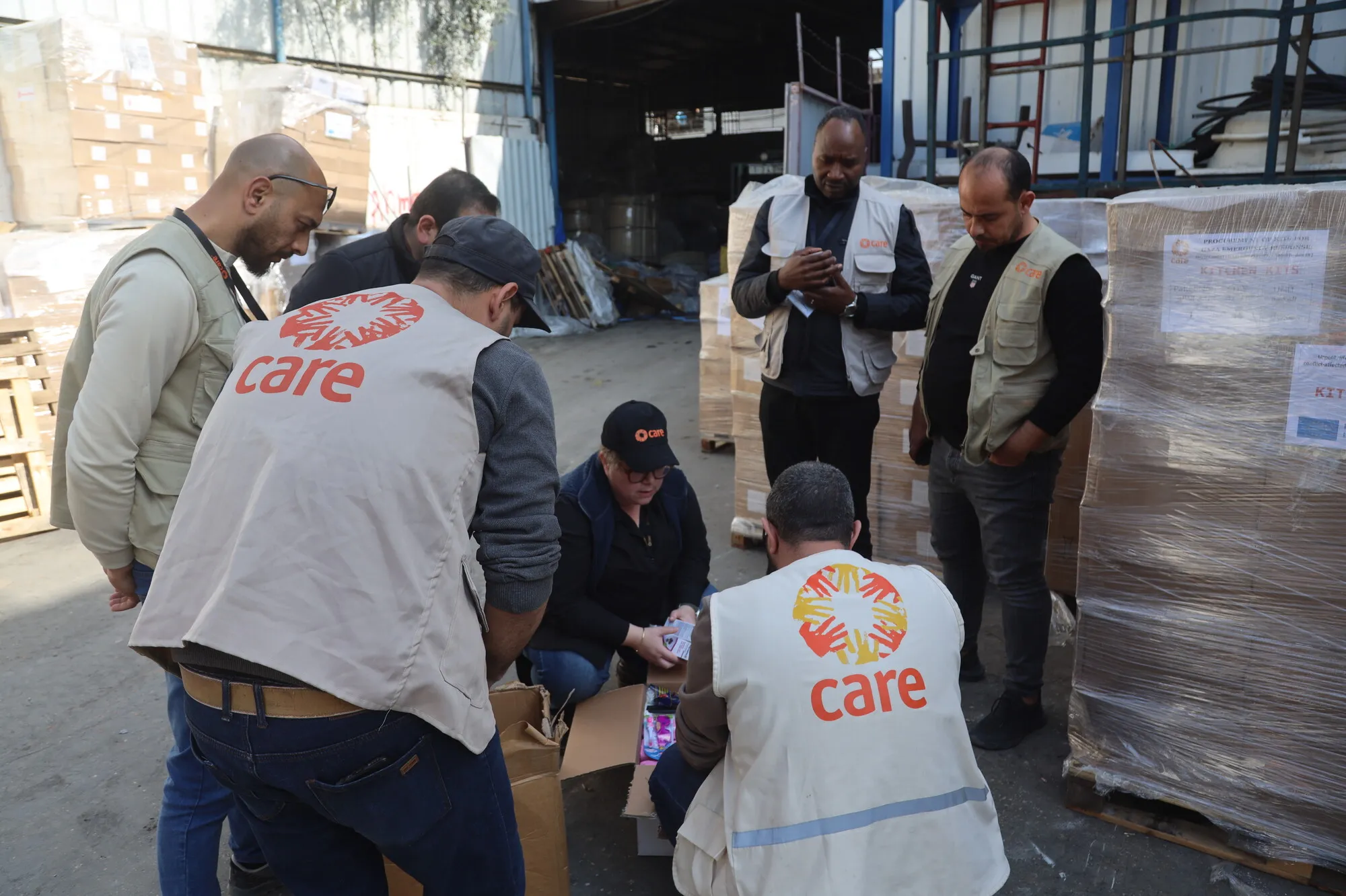 A group of six aid workers from CARE, wearing branded vests, gather outside a warehouse, examining supplies that were unpacked from a box on the ground.