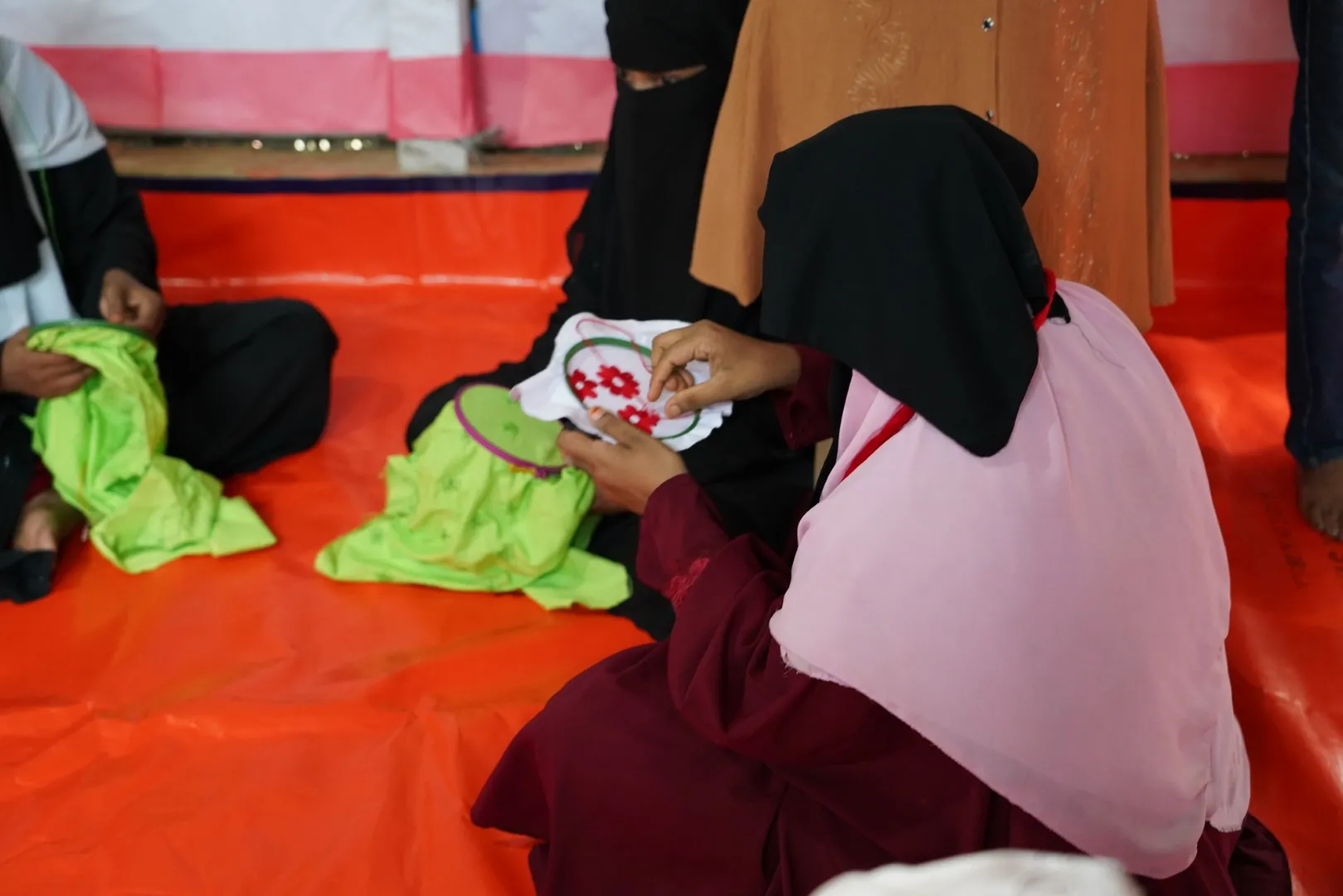 A close-up, over-the-shoulder photo shows several women seated on a bright orange floor mat, one of whom is focused on embroidering a floral pattern onto a white fabric.