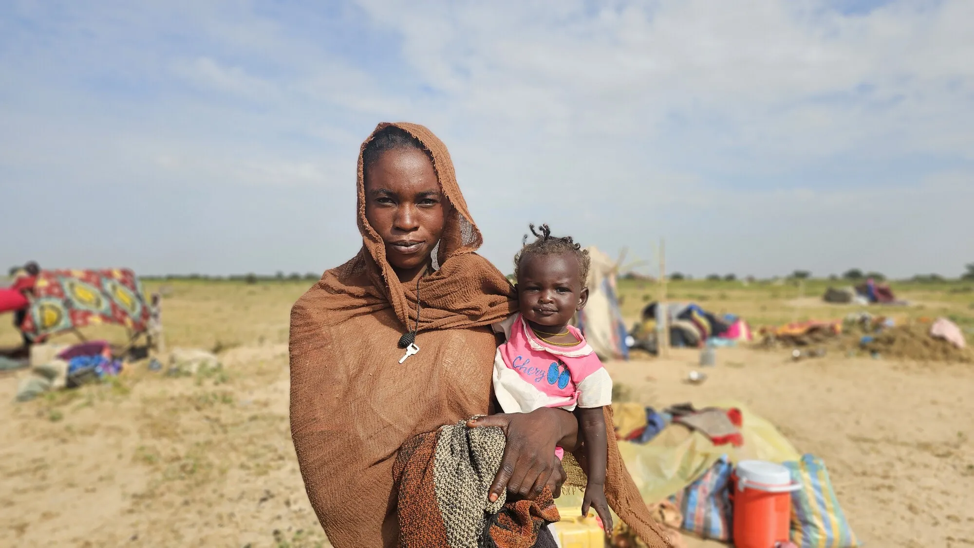 A portrait of a mother from Sudan holding her smiling baby in a dusty field with a makeshift shelter in the background.