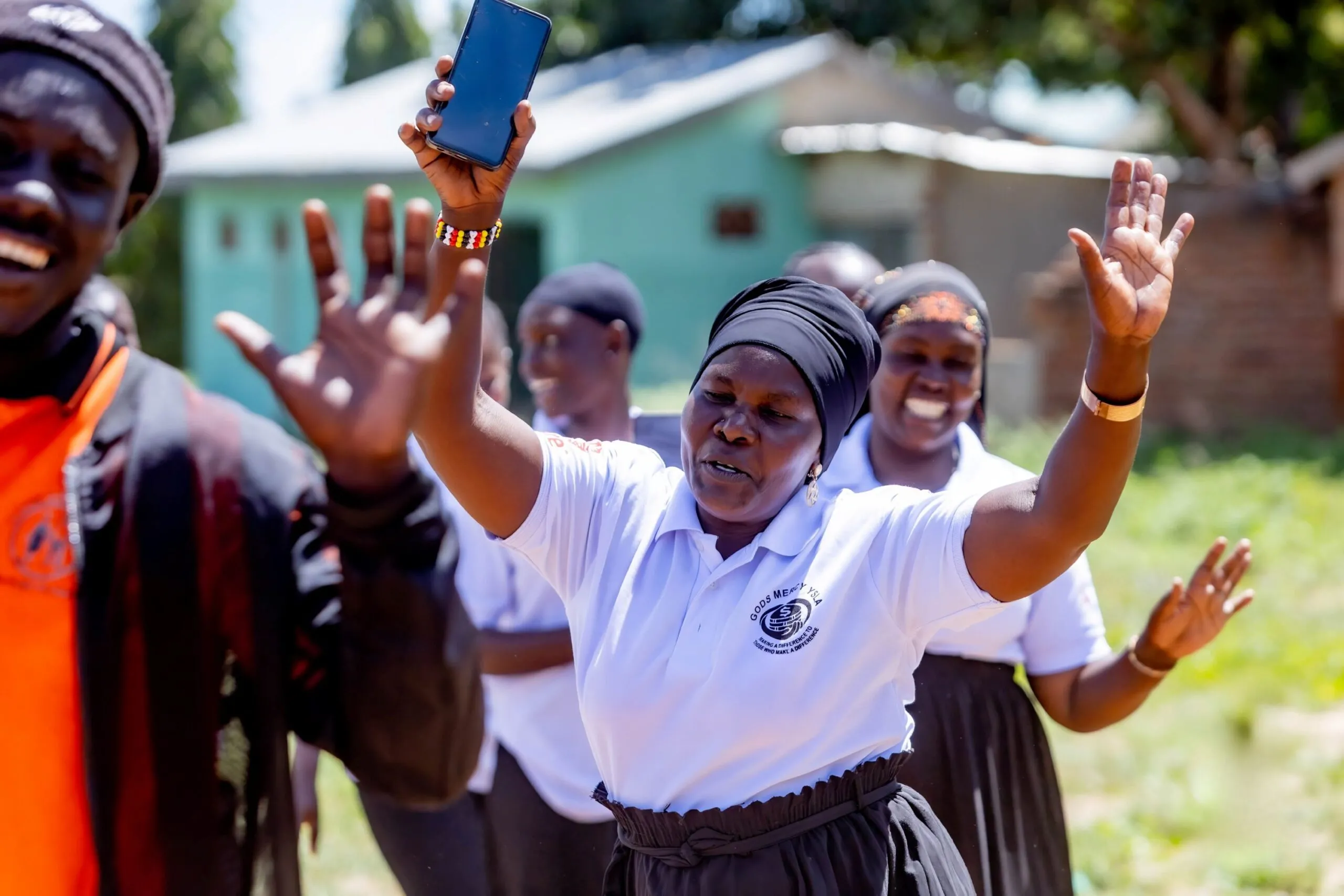 A joyous woman in Uganda, a Village Savings and Loan Association or VSLA member, raises her hands and holds up a mobile phone, celebrating with a group of other women and men, also VSLA members, outdoors in a village setting.
