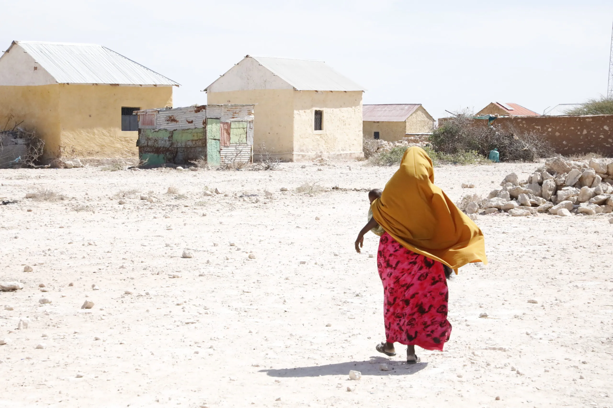 A Somali woman carrying a small child walks away from the camera across a dry, dusty landscape. In the background, several pale yellow buildings with tin roofs stand under a bright, hazy sky.