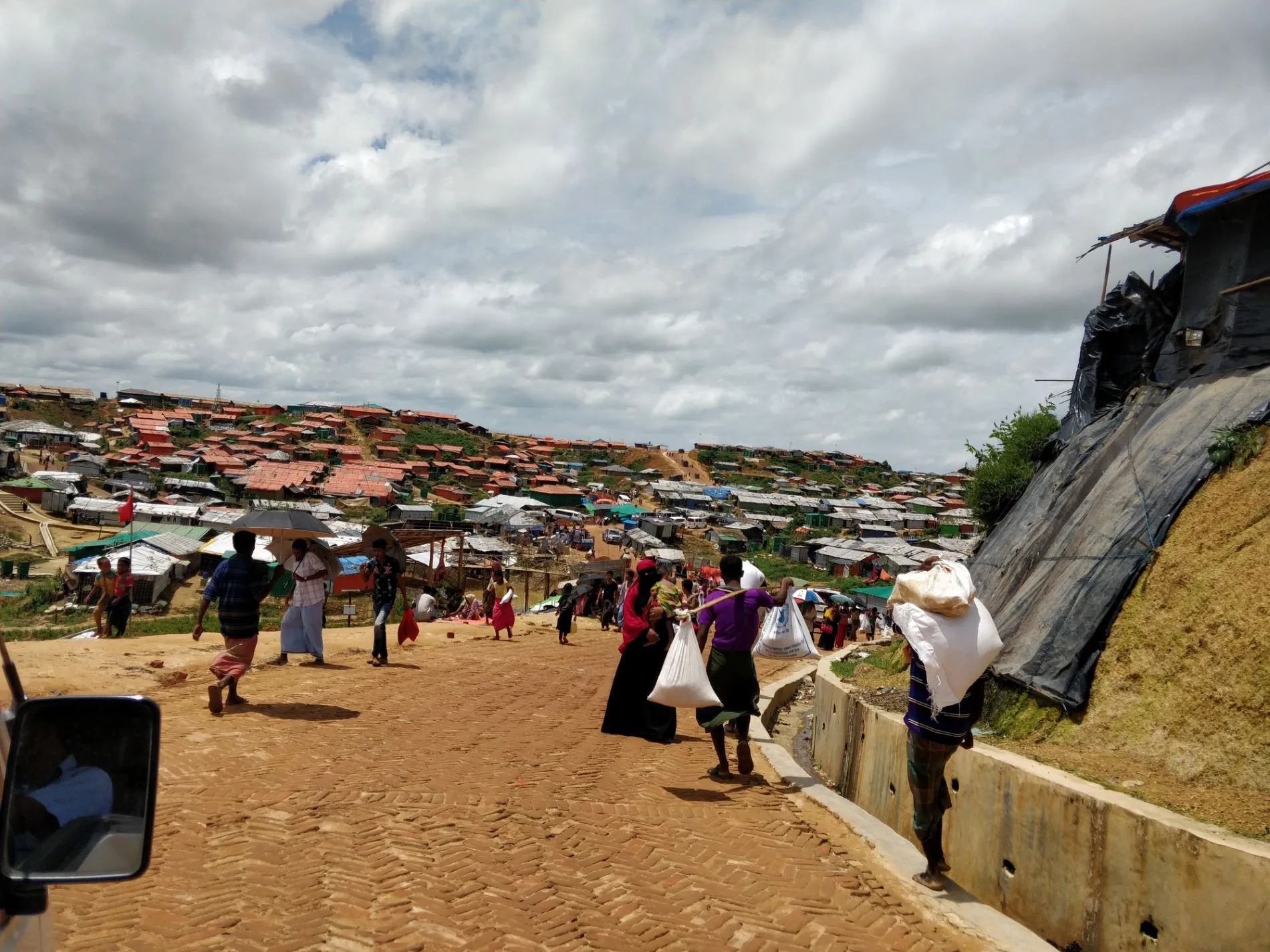 A low-angle photo shows a bustling camp road with people walking and carrying supplies. The camp's makeshift shelters climb a steep hill under a cloudy sky. A side mirror from a vehicle is visible in the foreground.