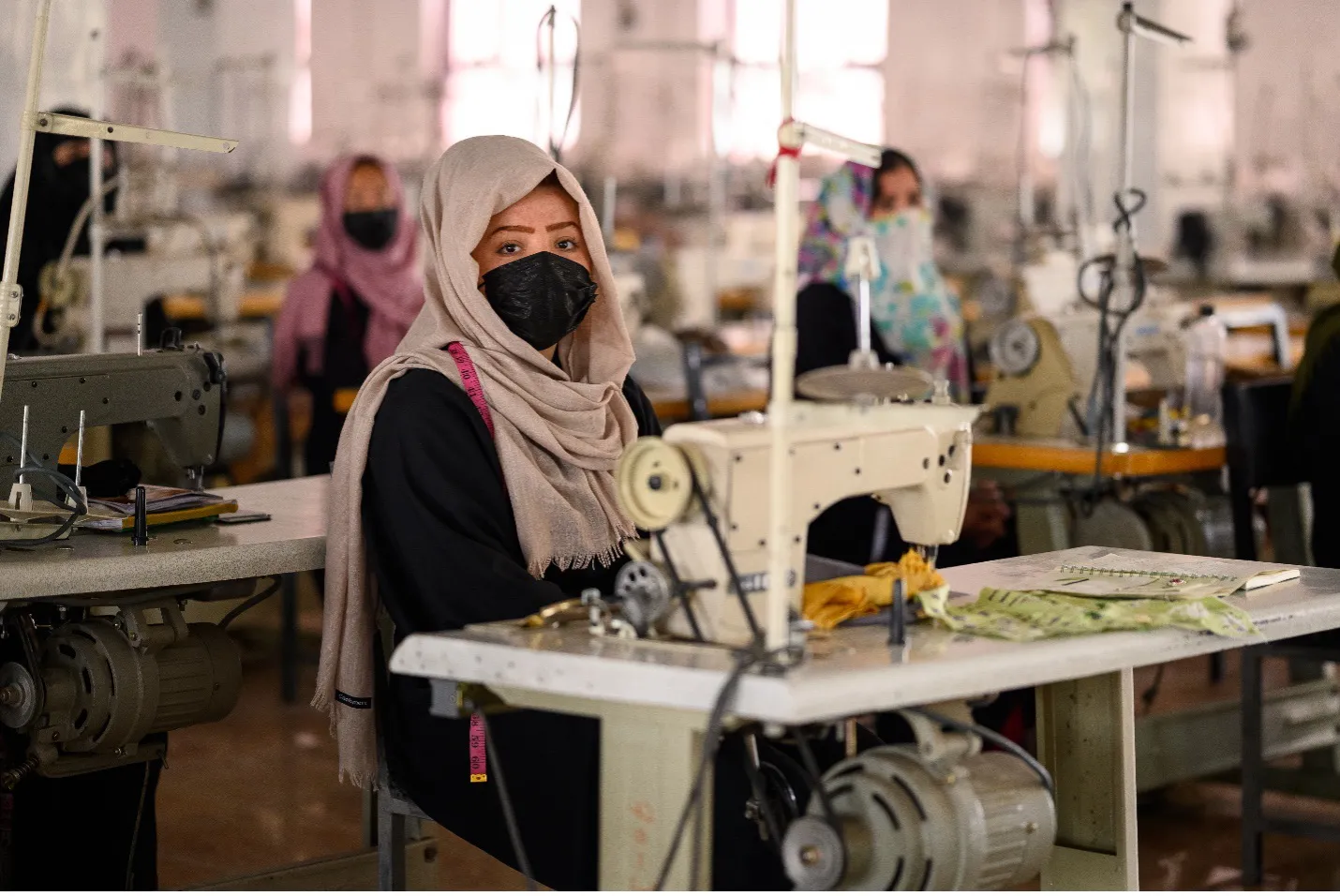 Portrait of a woman with a head covering and mask at a sewing machine looking directly at the camera. Other women are in the background.