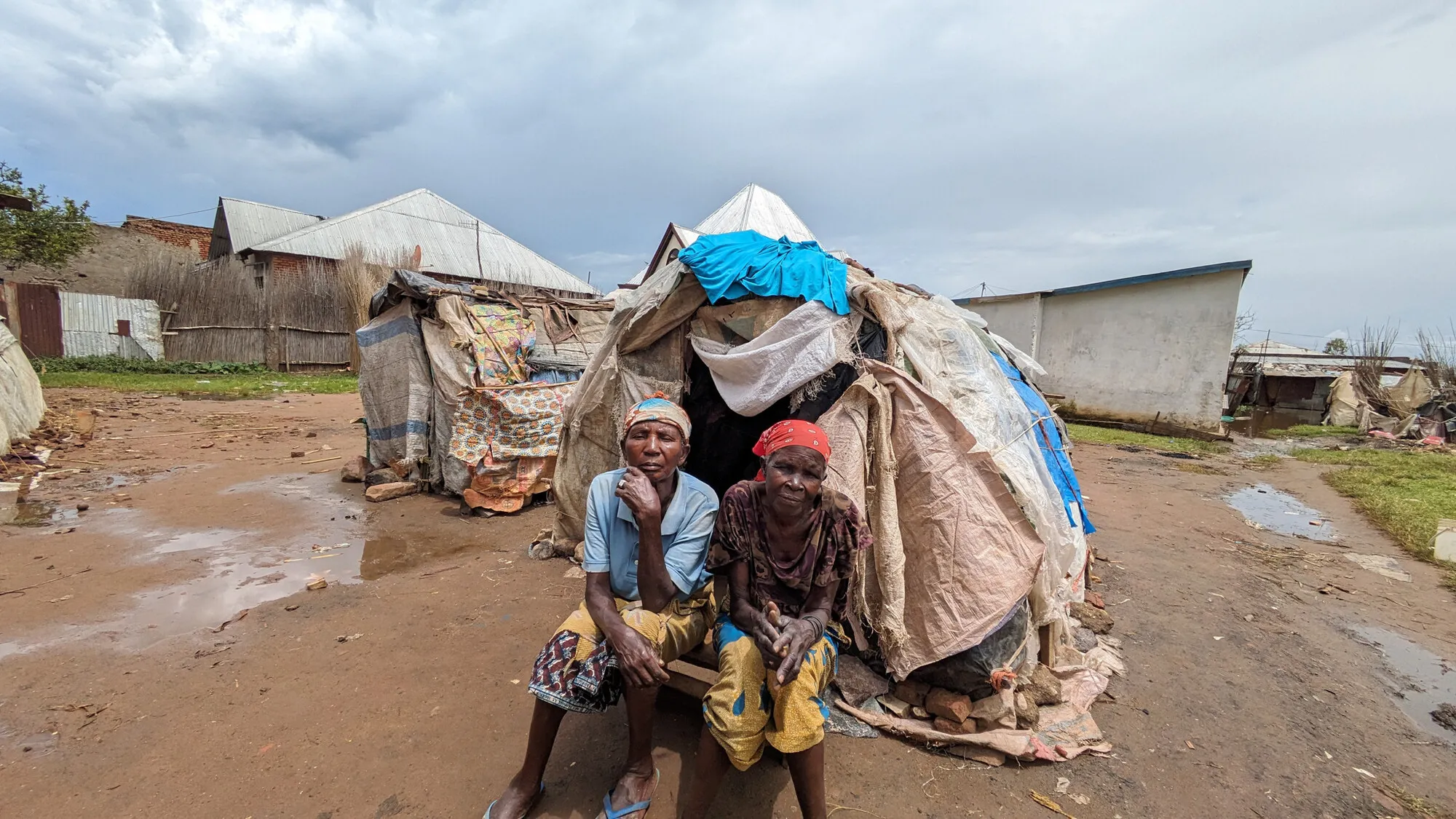 Two women sit outside a makeshift hut constructed from plastic sheet and bamboo.