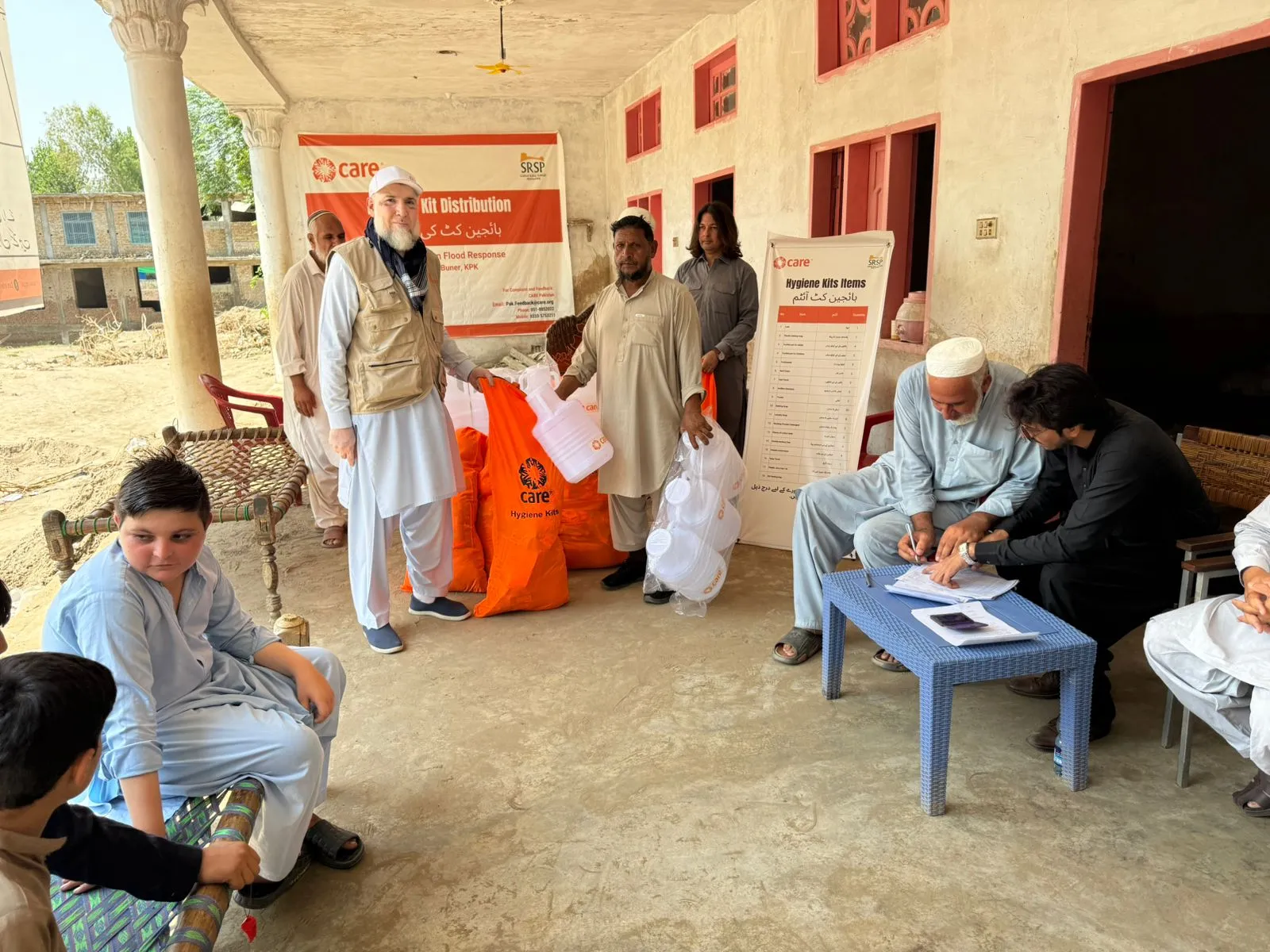 Pakistani men and boys gather under a porch for a flood relief distribution, with two men signing documents at a table. Orange 