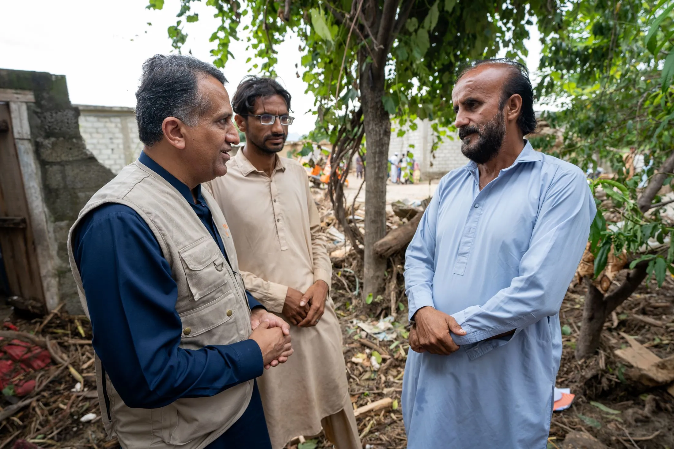 A photo shows three men standing and talking outdoors, surrounded by debris. The man on the left, who appears to be the CARE Pakistan country director, wears a dark shirt and a tan emergency vest. The man in the middle wears a light brown tunic. The man on the right wears a light blue tunic.