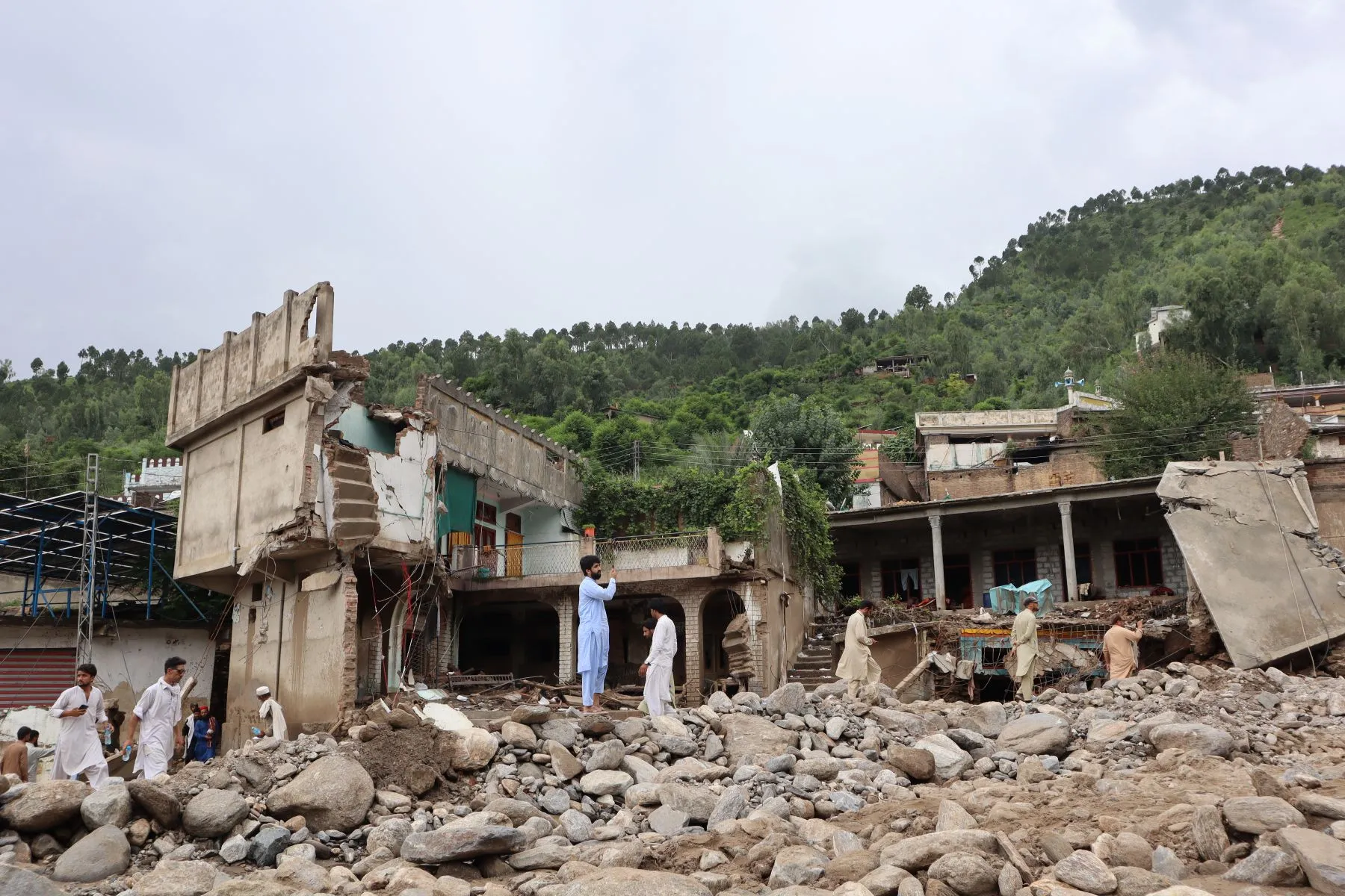 Men in traditional Pakistani clothing stand among rubble and large rocks, with a green hillside in the background, showing flood aftermath in a mountainous area.