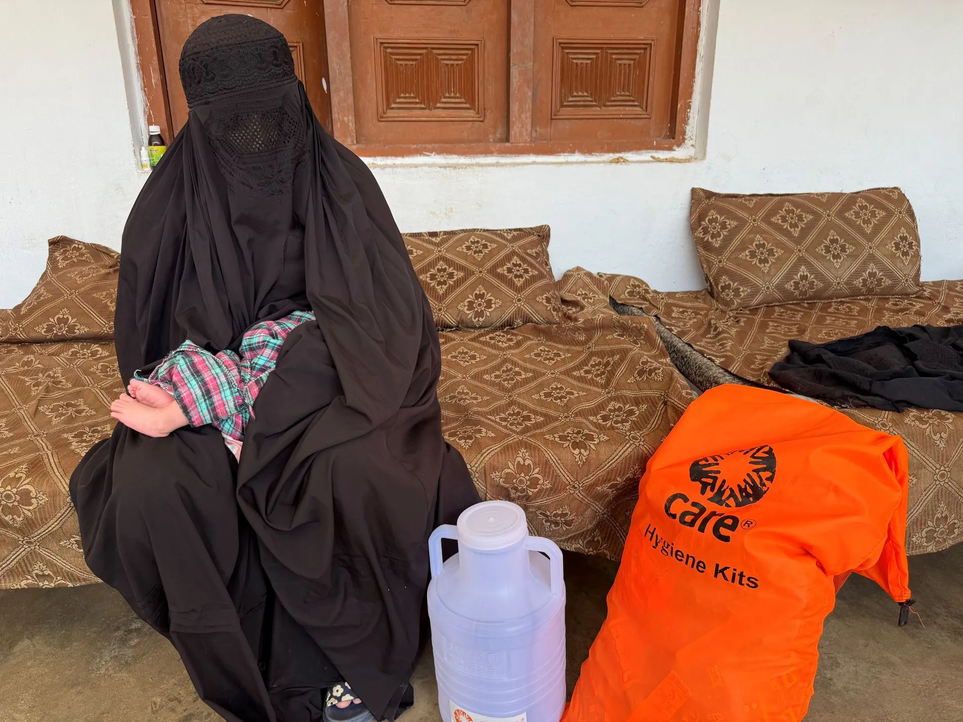 A Pakistani woman in a black burqa sits on a patterned divan, holding a baby wrapped in a plaid blanket. Nearby are a large white jug and a bright orange 
