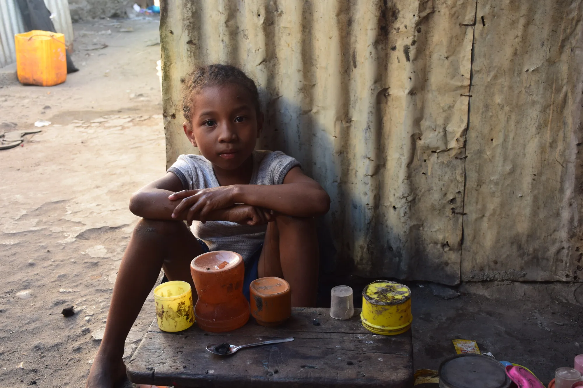 A child gazes at the camera while leaning against a corrugated iron (CI-sheet) wall.