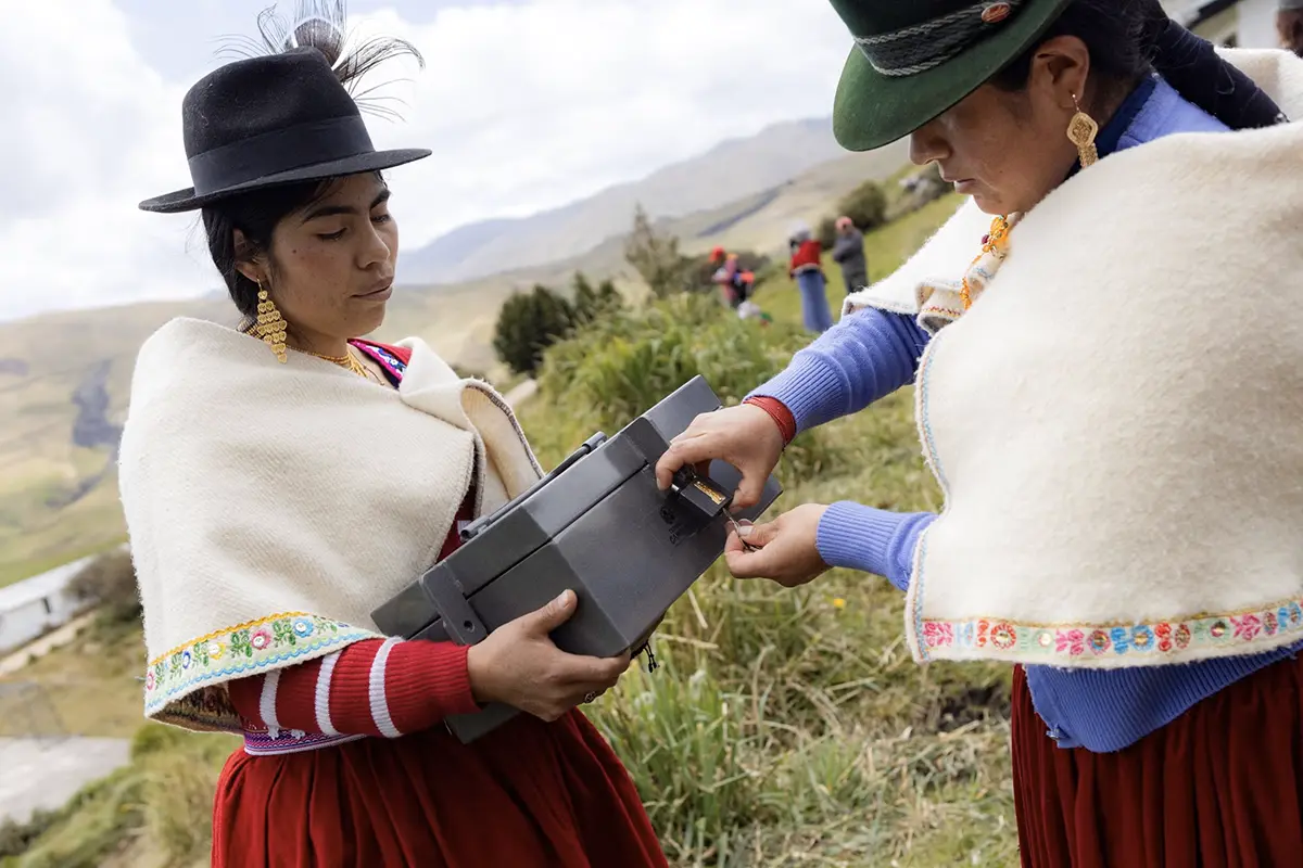 Two members of a Village Savings and Loan Association or VSLA in traditional Ecuadorian attire handling their group's metal savings box.