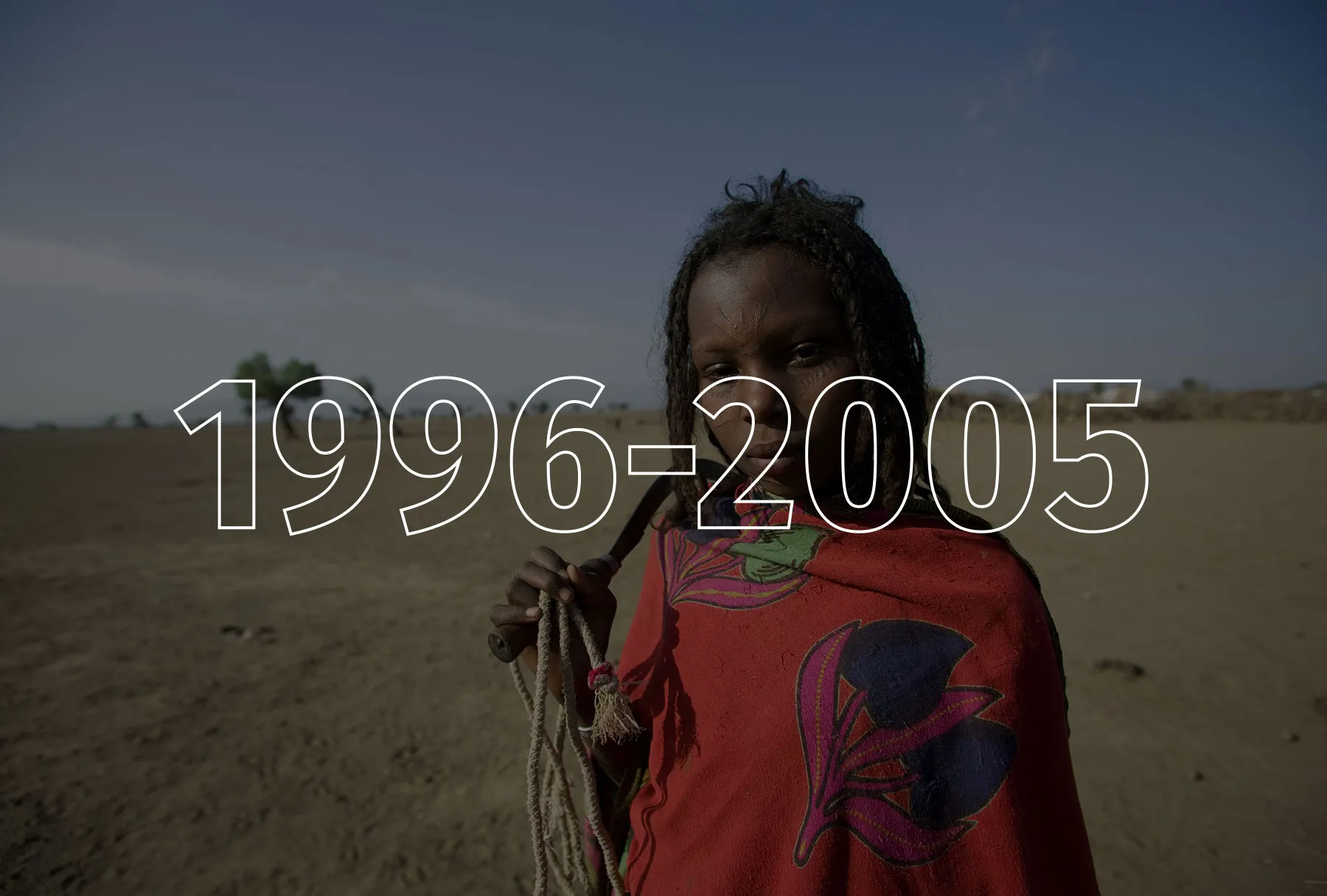 A girl wearing bright red and holding a thin rope and digging tool looks at the camera. Behind her is a wide expanse of dirt and sand with a couple of trees.