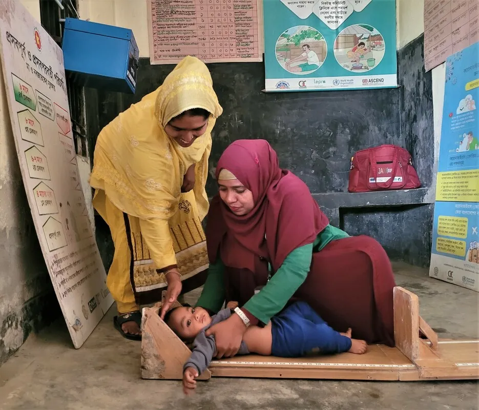 Two Bangladeshi women wearing head scarves bend over to measure a 7-month baby.