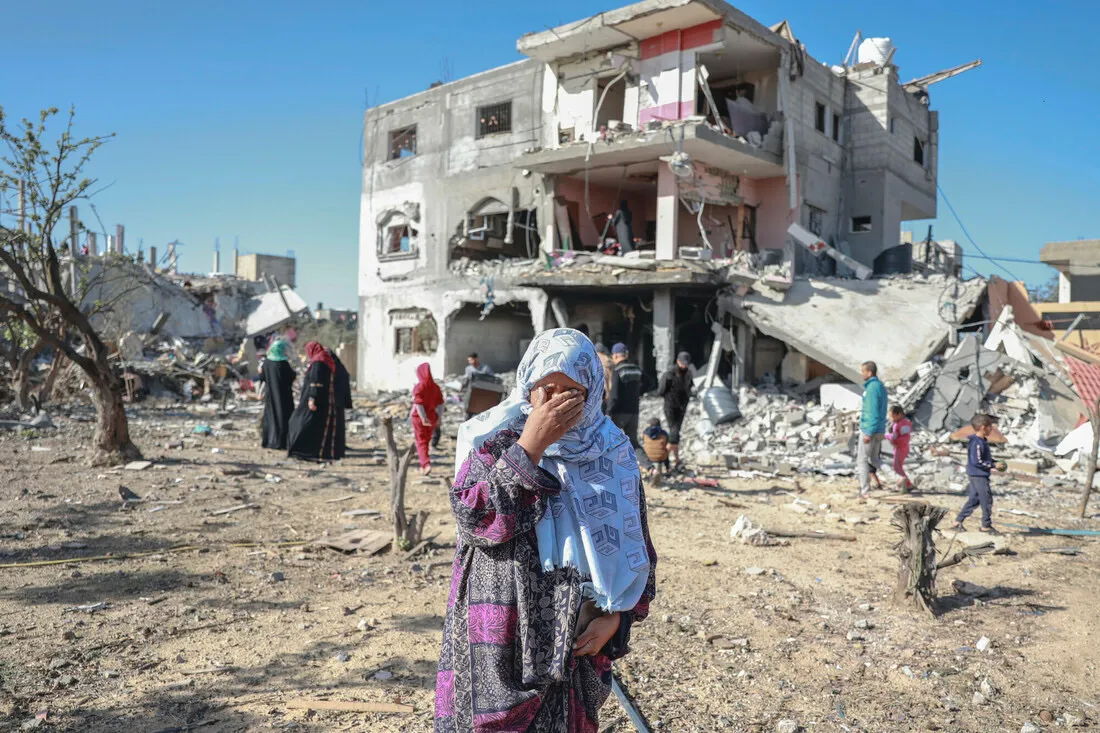 A woman wearing a headscarf covers her face with her hand. Behind her, people walk among rubble.
