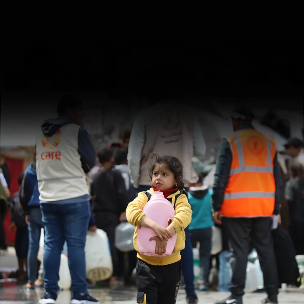 A young girl carries a jug of water. Behind her, a man wearing a CARE vest talks with other adults.