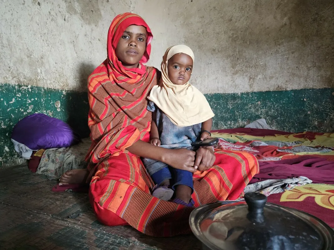 A woman sits in a small room and holds her young child in her lap.