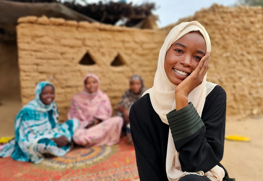 A young Sudanese woman smiles and rests her head in her hand. Her three sisters sit on a rug behind her.