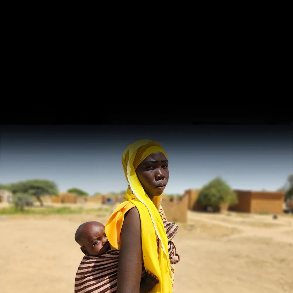 A woman stands in a dusty area with her young baby resting on her back.