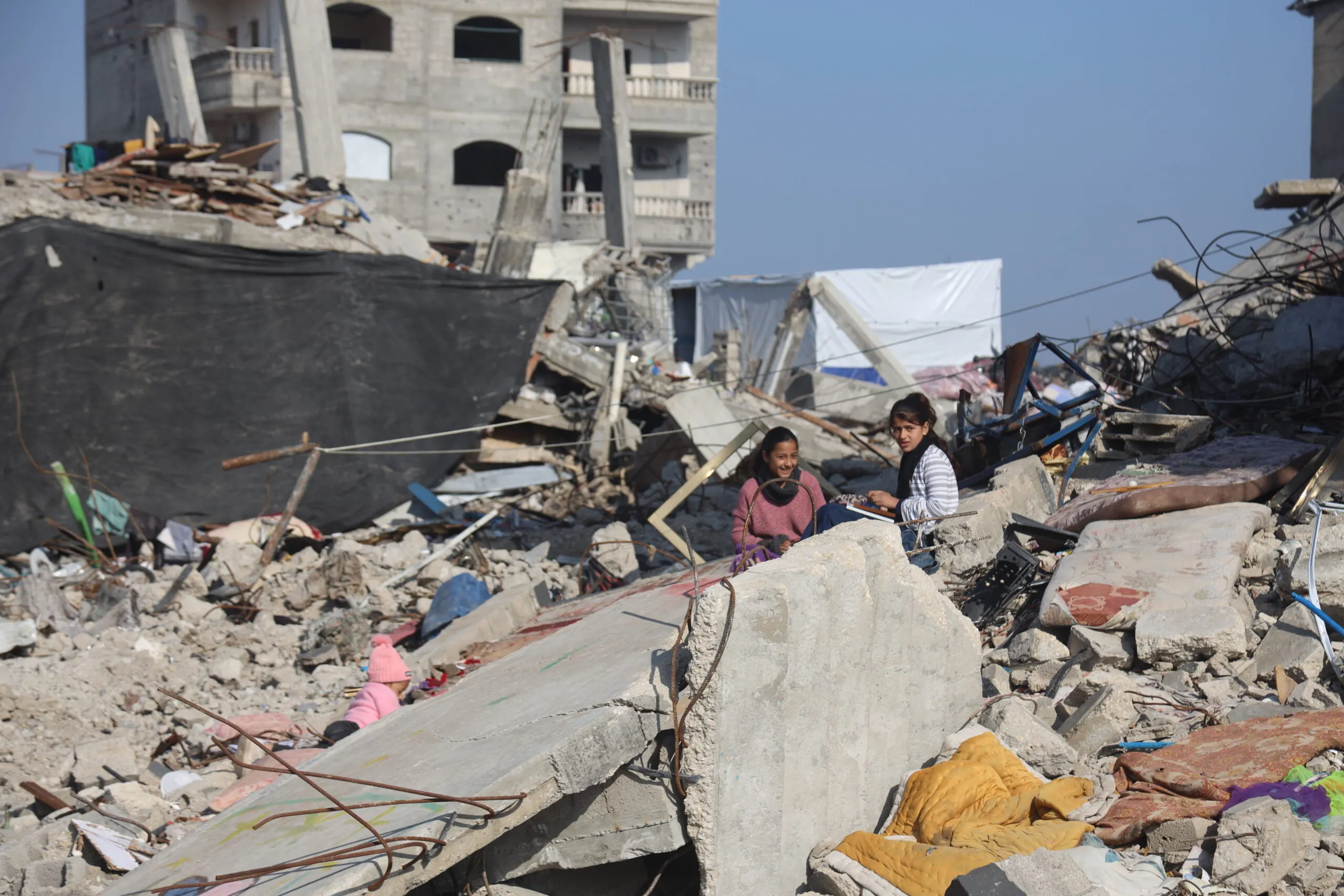 Two children sit atop slabs of broken, twisted concrete amidst rubble in Jabalia Camp, North Gaza.