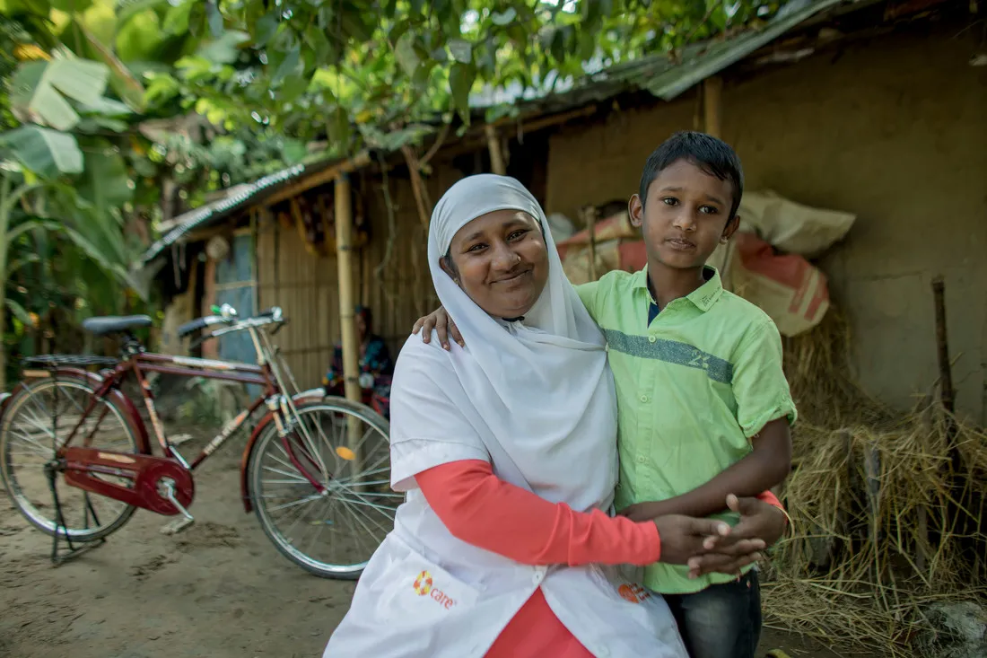 A woman sitting down smiles and hugs a young boy close to her. They are sitting in front of a small bamboo house with a bike parked outside.