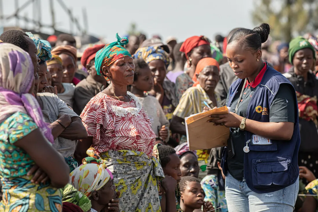 A Care staff member talking to a group of men, women, and children writing on paper.