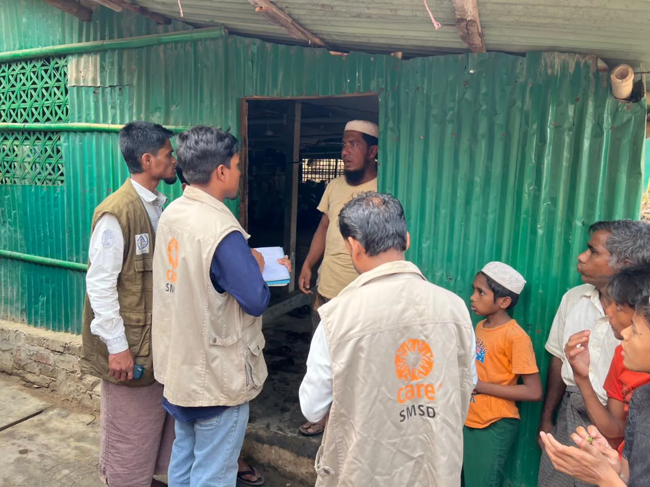 A group of people, including men wearing CARE vests, talk outside of a small green building.